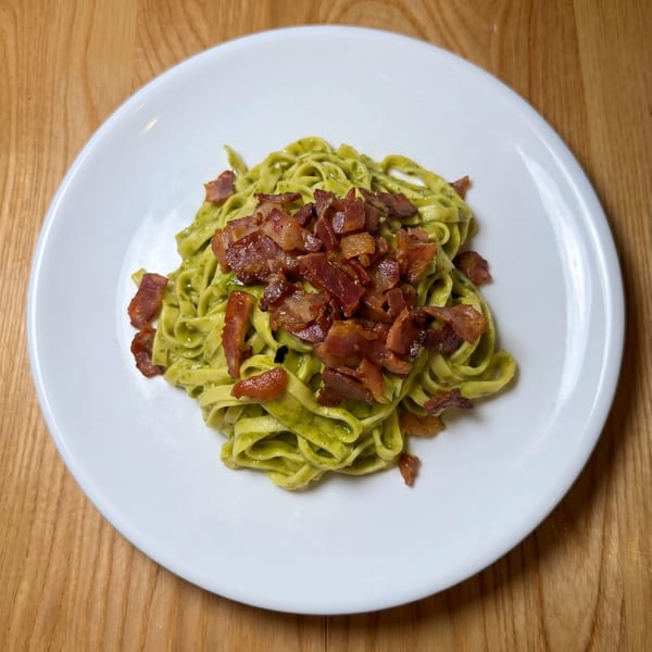 Green pesto tagliatelle pasta topped with crispy bacon bits on a white plate.